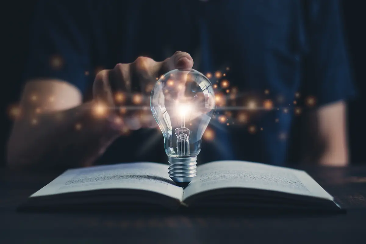 Glowing light bulb standing on an open book with a person seated in the shadowy background