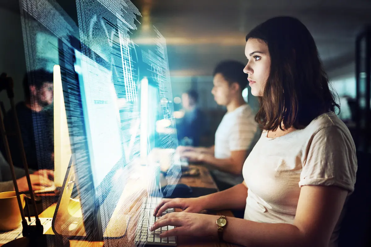 Young woman focused on computer screen displaying glowing blue code in a dim office with colleagues nearby