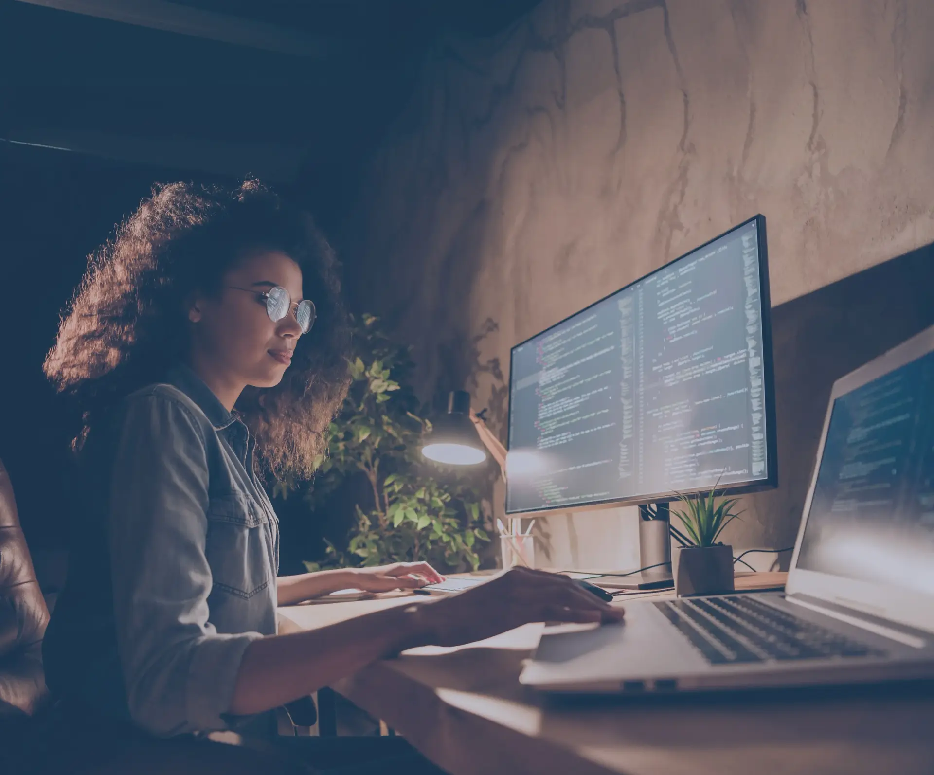 Young woman with curly hair and glasses coding on a monitor and laptop at night