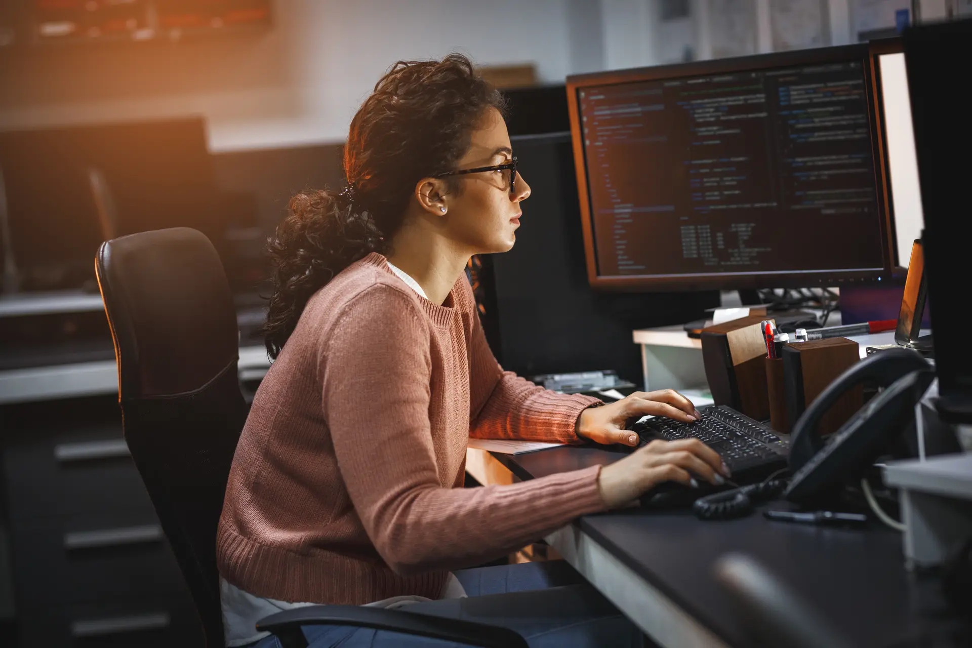 A woman wearing glasses and a pink sweater types on a keyboard while viewing code on a monitor.